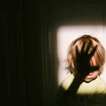 So rutscht die Hand nicht aus Anonymes kleines Kind, das in einem quadratischen Lichtfleck neben einer Tür in einer alten Wohnung sitzt, mit dem Schatten einer Erwachsenenhand über seinem Gesicht.