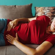 Welche Ernährung hilft bei einem Reizdarm? Eine Frau liegt mit Schmerzen im Bauch auf dem Sofa.