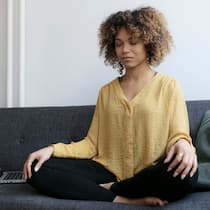 «Raus aus negativen Gedankenspiralen» Junge Frau sitzt mit Laptop auf dem Sofa und meditiert