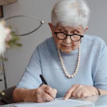 Prüfen Sie jetzt Testament und Erbvertrag Seniorin schreibt auf einem Papier und sitzt zuhause am Tisch