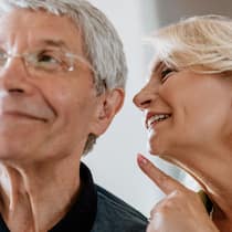 Mehr Lebensqualität im Alltag Ein älterer Mann mit Hörhilfe in seinem linken Ohr lächelt zufrieden vor sich hin während ihm seine Frau etwas ins Ohr flüstert.