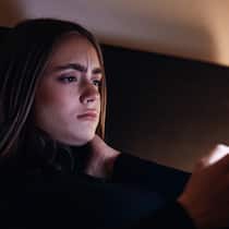 Wie bekommt man Ängste in den Griff? Eine junge Frau liegt mit besorgtem Gesicht im Bett und liest Nachrichten auf ihrem Handy