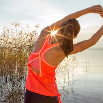6 Tricks, wie Sport schneller schlank macht Eine junge Frau dehnt sich vor dem Sport draussen in der Natur bei Sonnenaufgang