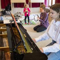 Між експлуататорами та бюрократизмом Eine junge Frau sitzt am Klavier und spielt ein Stück während im Hintergrund kleine Kinder spielen.