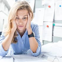 Macht uns die Arbeit krank? Eine Frau sitzt an ihrem Arbeitsplatz, der mit Akten überladen ist, stützt den Kopf in die Hände und macht ein sorgenvolles Gesicht.