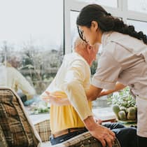 Warum Care-Arbeiterinnen kaum geschützt sind Eine Pflegerin hilft einem Senior beim Hinsetzen oder Aufstehen.