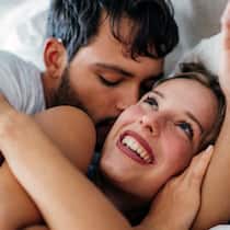 Tipps für mehr Spass im Bett Ein Paar liegt eng umschlungen im Bett, er küsst sie und sie lacht