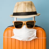 Bin ich im Sommer für Corona versichert? Ein kleiner oranger Reisekoffer ist mit einem Sonnenhut, Sonnenbrille und einer Corona-Schutzmaske drapiert.