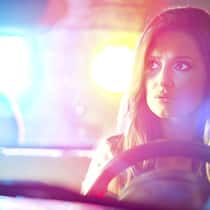 Was sind meine Rechte? Eine junge Frau sitzt am Steuer während hinter ihr die Blaulichtanlage eines Polizeiautos zu sehen ist. Diese Rechte haben Autolenkerinnen und Autolenker, wenn sie von der Polizei kontrolliert werden