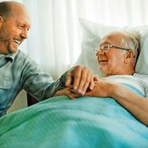 Wozu dient ein Vorsorgeauftrag? Ein älterer Mann liegt in einem Spitalbett und lächelt, ein zweiter Mann sitzt daneben und hält seine Hand. Für den Fall einer Urteilsunfähigkeit gibt es die Möglichkeit, eine Drittperson mit einem Vorsorgeauftrag zu bestimmen.