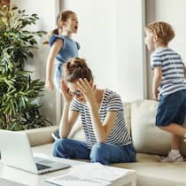 Ein Leben auf der Überholspur Müde junge Mutter sitzt auf dem Sofa und arbeitet am Laptop und an Dokumenten während kleine Kinder Spass haben und Krach machen