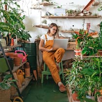 So macht die Krise Firmen innovativ Junge Frau sitzt in einer Gärtnerei auf einem Stuhl und arbeitet am Laptop.