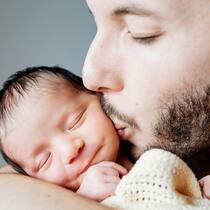 10 Regeln zum Papi-Urlaub Ein frischgebackener Vater hält sein neugeborenes Kind in den Armen und küsst es liebevoll auf die Wange.