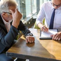 So hohe Zinsen von der Bank? Ein Bankangestellter zeichnet etwas auf ein Blatt Papier auf, um einen Kunden zu beraten, der leicht verzweifelt neben ihm sitzt.
