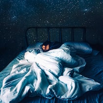 Was für einen gesunden Schlaf förderlich ist Eine Frau schläft tief in ihrem Bett. Im Hintergrund tut sich der Nachthimmel auf.
