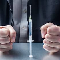 Darf der Chef beim Impfen und Testen mitreden? Ein Mann im Anzug sitzt an einem Tisch, vor ihm steht eine Spritze mit einem Covid-19-Impfstoff. Seine Hände sind zu Fäusten geballt.