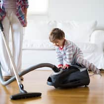 Der Ständerat bremst Frauen bei der Vorsorge aus A mother vacuums the floor of a bright living room while her young child playfully sits on the vacuum cleaner, creating a cheerful atmosphere in their home.