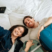 Wann der Chef die Ferien streichen darf Ein lachendes Paar liegt auf dem Bett im Hotelzimmer und macht ein Selfie.