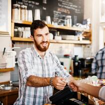 So funktioniert die Bundeshilfe für KMU und Selbständige Ein Restaurantbetreiber steht an der Kasse und nimmt Geld von einer Kundin