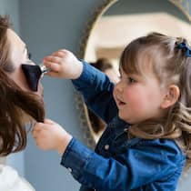 Muss ich trotz Kleinkind arbeiten? Eine Mutter wird von einem kleinen Kind mit einem Make-up Pinsel geschminkt.