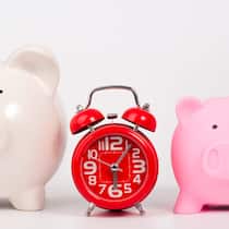 Werden meine Ergänzungsleistungen gekürzt? Ein grosses weisses Sparschwein steht links, ein kleineres rosa Sparschwein rechts und in der Mitte ist eine Tischuhr platziert.