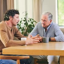 Ist ein Darlehen der Eltern das Beste? Ein erwachsener Sohn und sein Vater sitzen am Tisch währen der Sohn mit den Händen gestikulierend seine finanzielle Situation dem Vater schildert.
