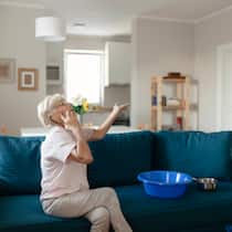 Wann gibts Geld vom Vermieter? Ältere Dame sitzt auf dem Sofa. Auf dem Sofa liegen Eimer. Sie telefoniert und schaut an die Decke hoch.