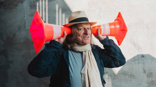 Andres Bosshard hält sich zwei orange Warnkegel an die Ohren wie alte Hörgeräte - Andres Bosshard sieht mit den Ohren. Der 70-jährige Musiker veranstaltet Klangspaziergänge und berät Behörden. Sein Lieblingsgeräusch? Menschengemurmel