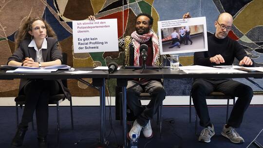 Mohamed Wa Baile an der Medienkonferenz zum neuen Urteil des EGMR. Er hält Plakate hoch, neben ihm zwei Juristinnen.