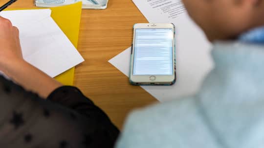 Ein Smartphone liegt zwischen Zetteln auf einem Schultisch.