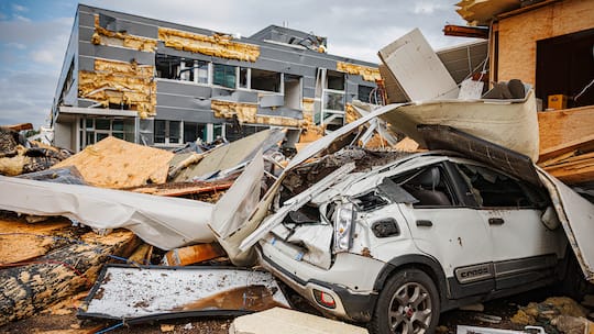 Zu sehen ist ein Gebäude, das von einem Sturm verwüstet wurde, die Fassade ist teilweise abgerissen. Davor sieht man ein zusammengebrochenes Gebäude und darunter begraben ein zerbeultes, weisses Auto.
