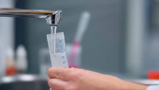 Nahaufnahme der Hand einer Biologin, die Leitungswasser in einem Reagenzglas auffängt, um eine Wasserprobe im Labor zu untersuchen.