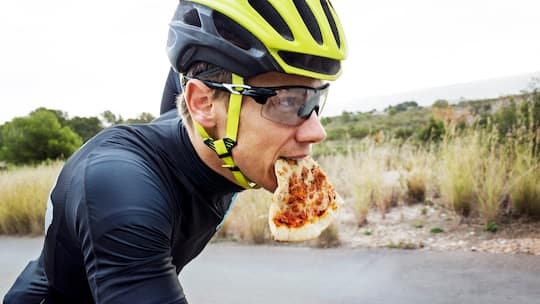 Ein Velofahrer mit Helm und Brille hat ein Stück Pizza im Mund. Die wichtigsten Fragen zu Ernährung und Sport.