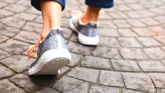 Ein Mensch geht über Pflastersteine. Man sieht nur die Füsse von hinten. Sie stecken in Turnschuhen