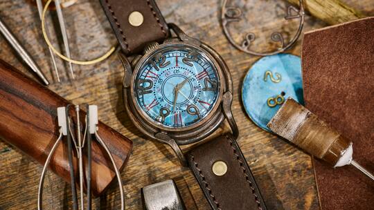 Eine Uhr mit braunem Lederarmband und türkis-farbigen Zifferblatt liegt auf einem Holztisch mit Utensilien eines Uhrmachers.