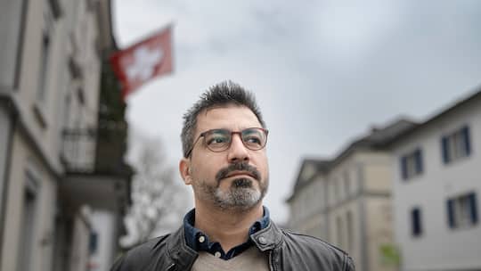 Ein Mann mit Brille und kleinem Bart schaut an der Kamera vorbei, hinter ihm Häuser und eine Schweizerfahne.