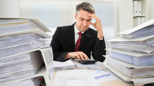 Mann im Anzug sitzt in Mitten einer Menge gestapelten Ordnern und rechnet auf dem Taschenrechner etwas aus.