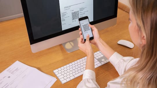 Eine Frau sitzt mit ihrem Smartphone vor einem Computerbildschirm und scannt mit ihrer Handykamera den abgebildeten QR-Code.