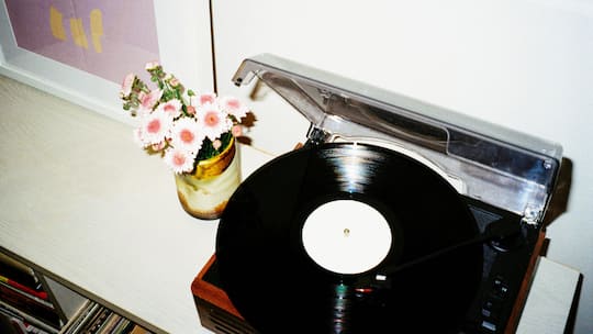 Ein Retro-Plattenspieler auf einem weißen Regal, aufgenommen vor einer weißen Wand. Neben dem Plattenspieler stehen eine kleine Vase mit rosa Blumen und ein gerahmtes Bild. Unter dem Regal sind Schallplatten und Bücher zu sehen.