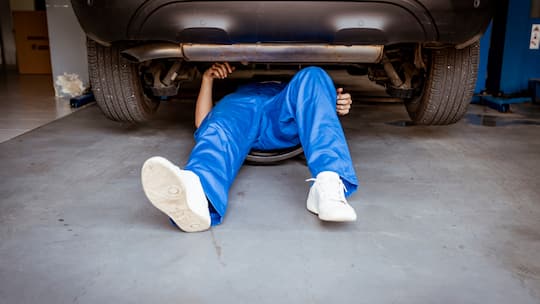 Ein Automechaniker in blauer Arbeitsuniform liegt unter einem Auto, das er gerade am Reparieren ist. Bei Offerten zu Autoreparaturen ist es wichtig, mehrere zu vergleichen, selbst wenn man wegen der Autoversicherung an eine Partnergarage gebunden ist.