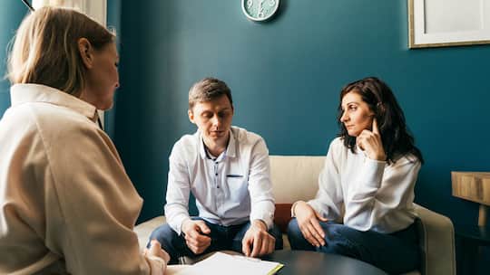 Ein junges Paar sitzt in einem Büroraum zusammen mit einer Beraterin. Die Frau wirft dem Mann einen kühlen Blick zu. Ist ein Scheidungsverfahren hängig, fallen gegenseitige Begünstigungen aus Testament oder Erbvertrag von Gesetzes wegen dahin.