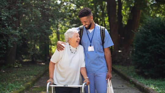 Ein Pfleger eines Heims geht auf einem kleinen Waldstück zusammen mit einer Heimbewohnerin und nimmt sie herzlich in den Arm.