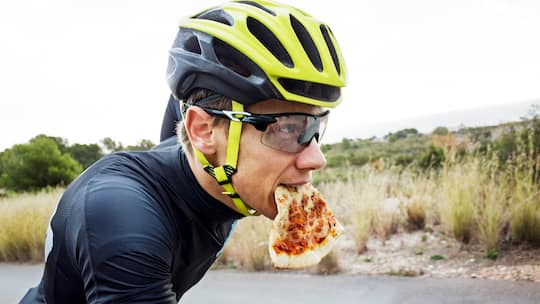 Ein Velofahrer mit Helm und Brille hat ein Stück Pizza im Mund. Die wichtigsten Fragen zu Ernährung und Sport.