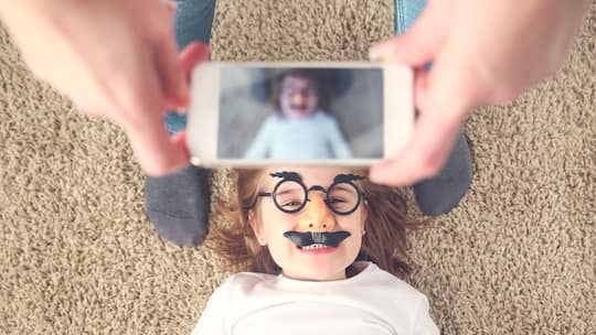 Kleines Mädchen mit Brille und Schnurrbart verkleidet liegt auf dem Teppich und wir mit dem Smartphone fotografiert