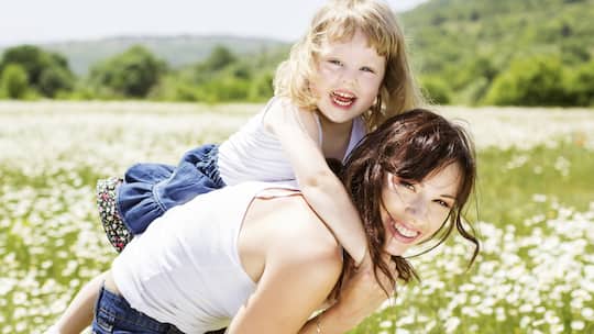 Eine Mutter trägt ihre kleine Tochter auf den Schultern und steht dabei auf einer blühenden Wiese.