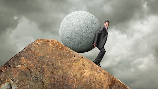 Ein Geschäftsmann versucht einen grossen kugelförmigen Stein über einen felsigen Gipfel zu stossen.