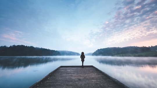 Eine Frau steht in der Ferne auf einem Holzsteg an einem See.