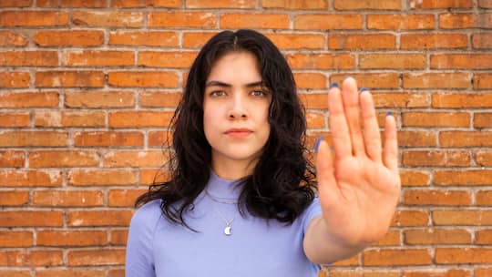 Eine junge Frau steht vor einer Mauer und streckt ihre Hand aus, mit der Handfläche Richtung Kamera.