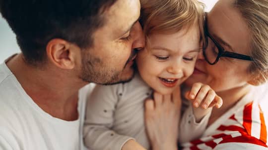 Mann und Frau küssen ein Kind - Eltern möchten für ihre Kinder da sein, daran ist nichts verkehrt. Ein neuer Elterntyp übertreibt es jedoch dermassen mit der Fürsorge, dass den Kindern Schaden entsteht. Die Rede ist von Klettverschluss-Eltern