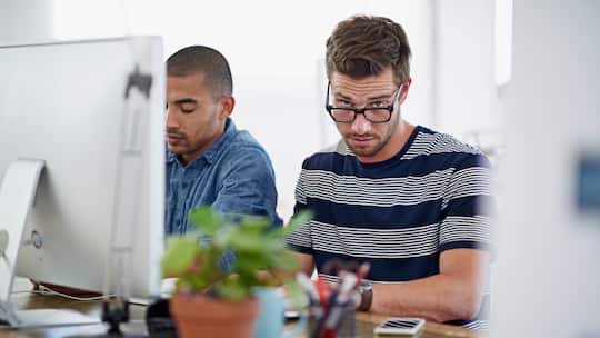 Ein Mitarbeiter sitzt vor dem Computer und blickt etwas genervt über seine Brille hinweg in die Kamera.